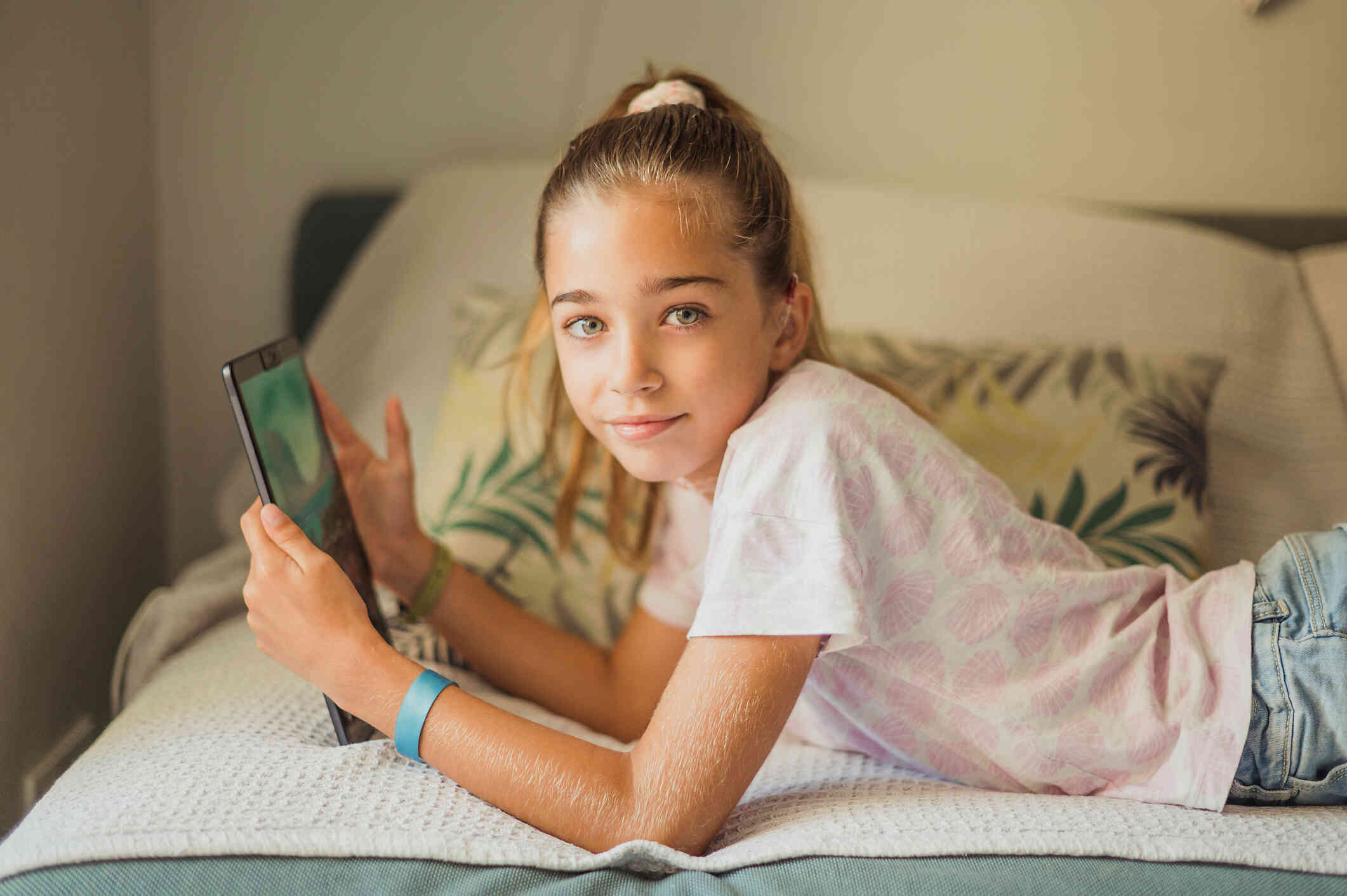 A young girl with a ponytail lays on her stomach on her bed with her tablet in her hands as she looks at the camera.