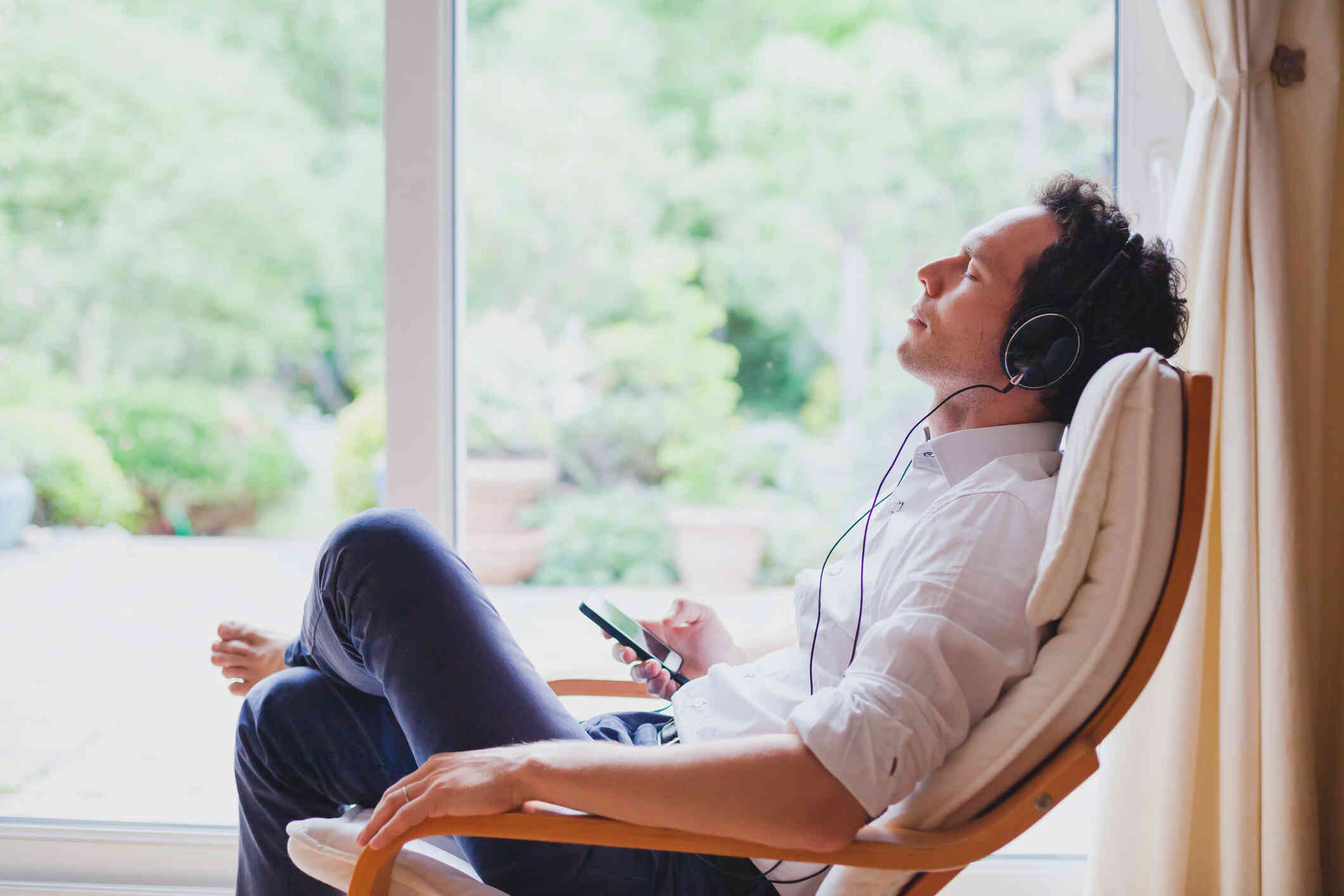 A man sits reclined in a chair with his eyes closed while wearing headphones and holding his phone in his hand.