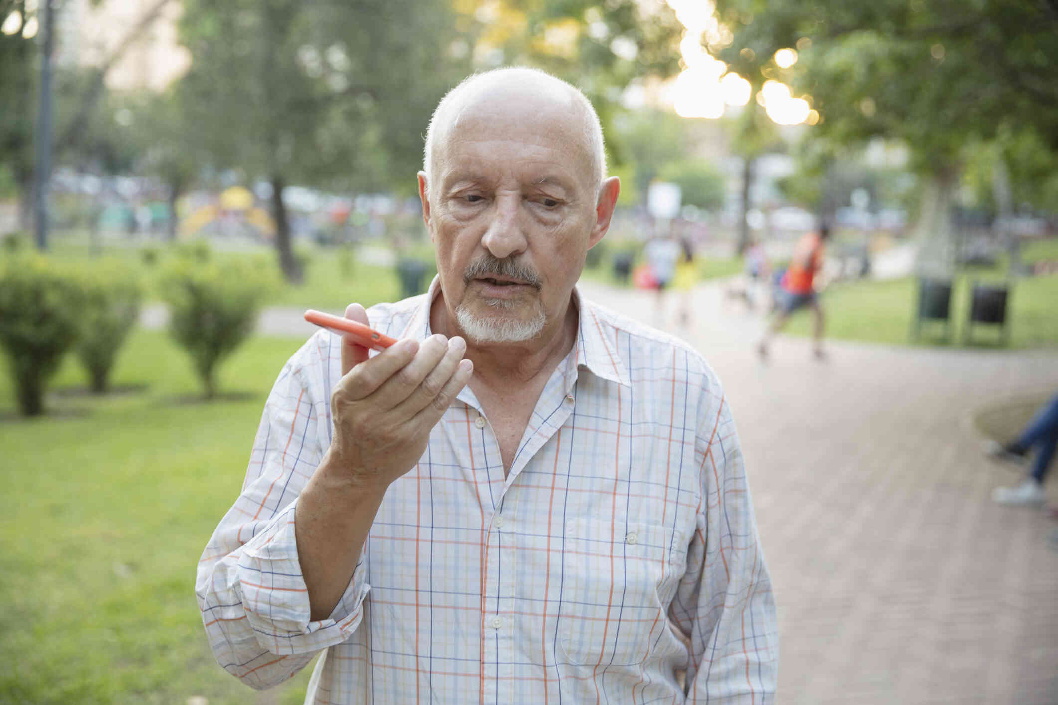 A mature man in a plaid shirts walks outside on a sunny day and talks on the phone in his hand with the phone on speakerphone.