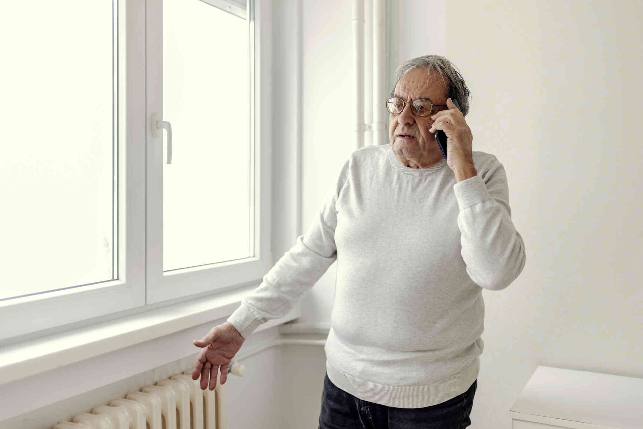 An elderly man in a white shirt stands in his home and talks on the phone with a worried expression.