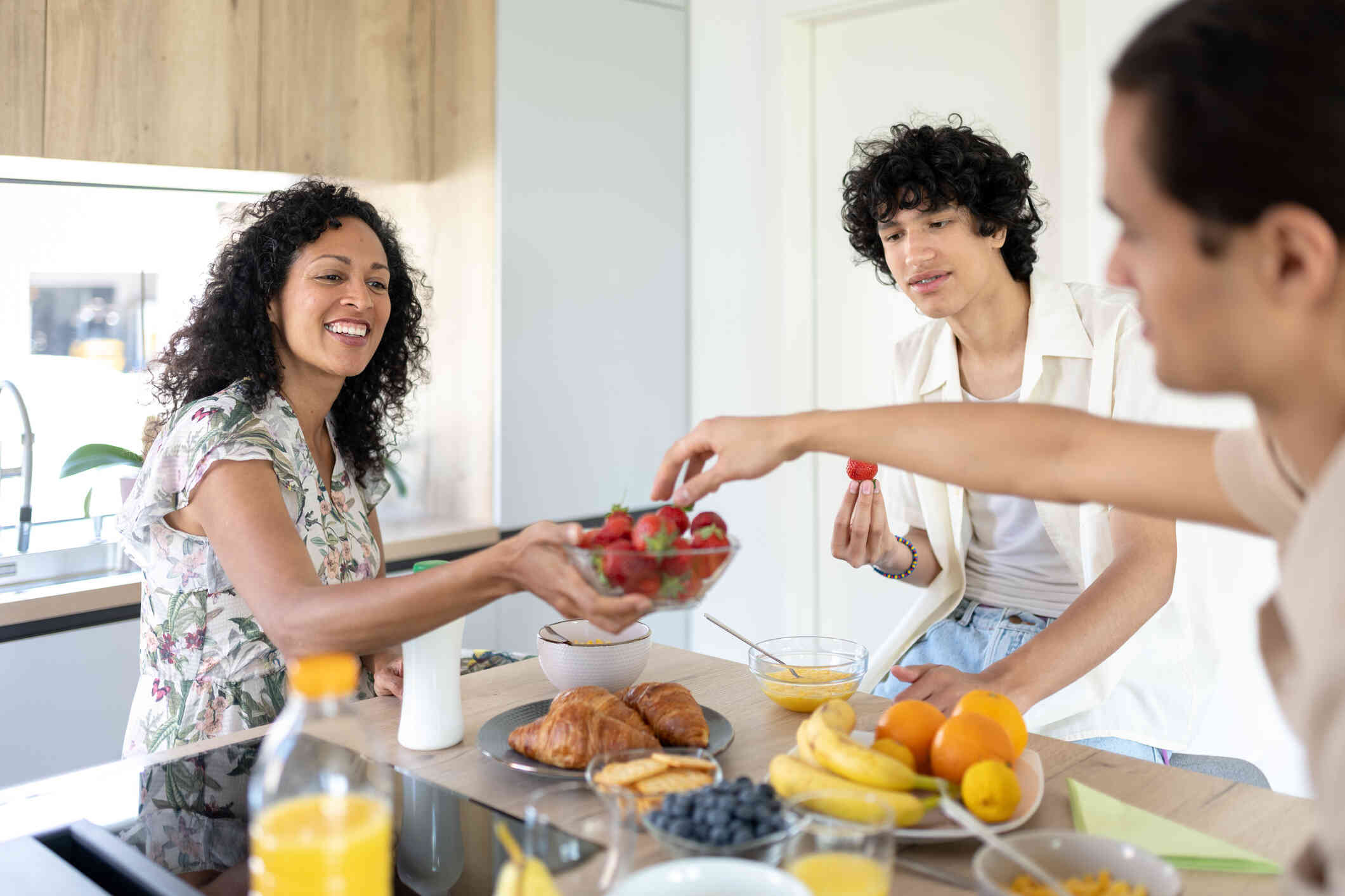 A mother, father, and teen son gather around the kitchen counter for breakfast as the dad passes the mother a bowl of strawberries.