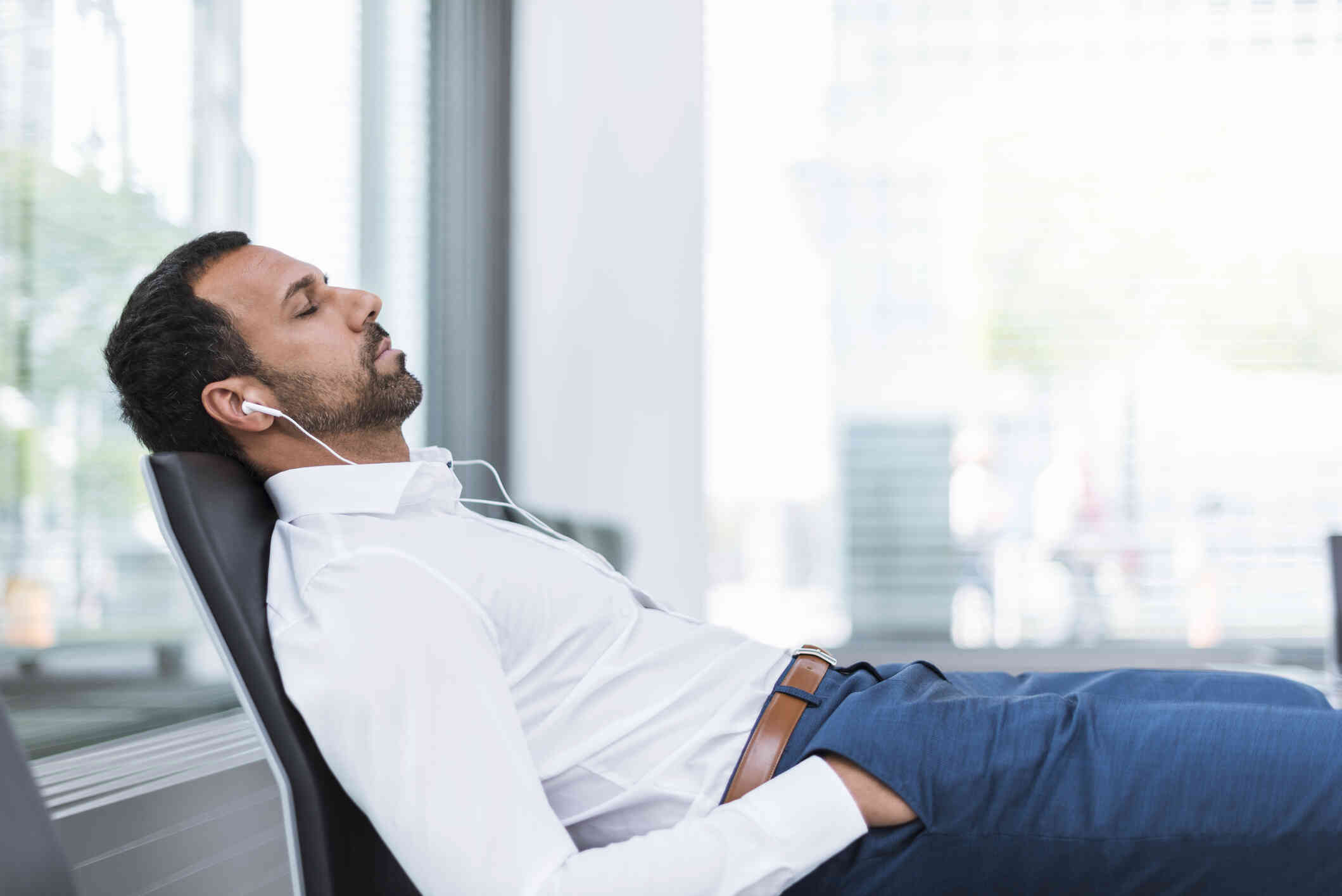 A man in a white button down shirt reclines in his office chair and closes his eyes while wearing headphones.