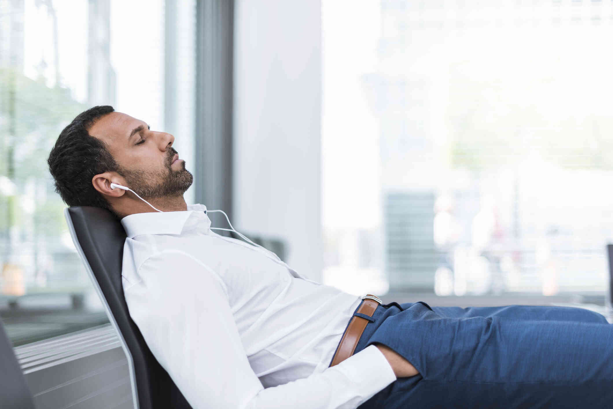 A middle aged man in a white button down shirt reclines in his chair while wearing white earphones and closing his eyes.