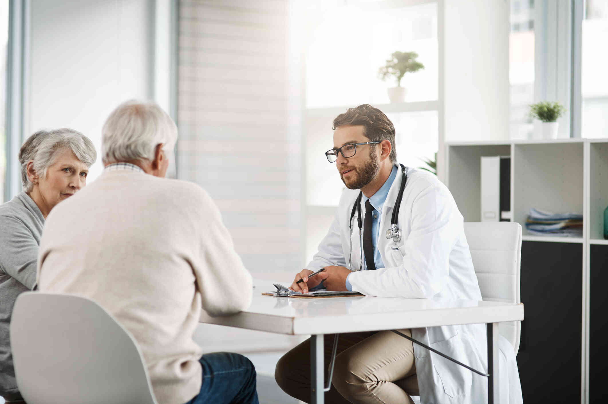 A alderly man and woman sit at a white table in the doctors office across from their male doctor.