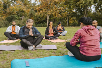 A group of men and women sit outside on yoga mats with their legs crossed and their hands against their chest in prayer position.