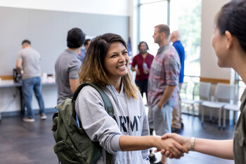 A woman wearing an army hoodie and a backpack smiles as she stands in a room with other people and shakes someone's hand.