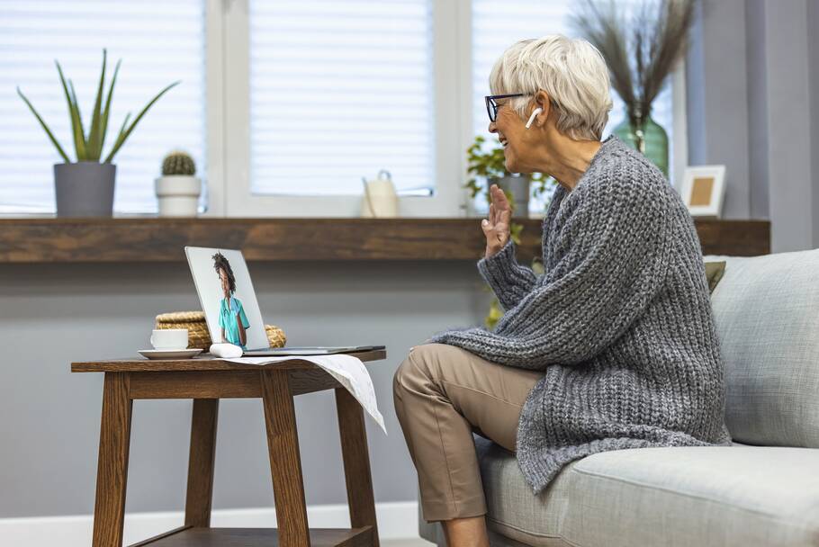 An elderly woman seated on her couch waves and smiles at her laptop during a video call.