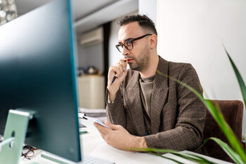 A man wearing glasses and a brown blazer sits at a desk and looks at a desktop computer while chewing on the end of a pen in his hand.