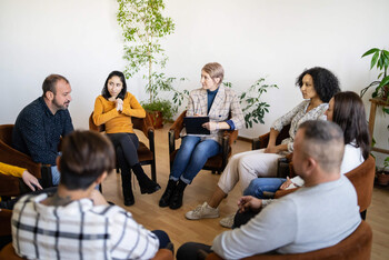 A group of people sit in a circle and look at a man who is speaking to the group.