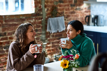 Two women in sweaters smile at each other as they sit at a table holding coffee mugs.