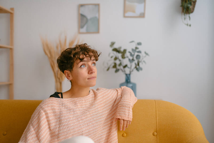 A woman in a striped sweater sits on a yellow couch and gazes off while deep in thought.