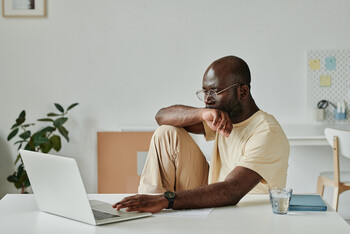 A man wearing glasses and a yellow t-shirt sits at a desk with his legs curled up and looks at a laptop screen.