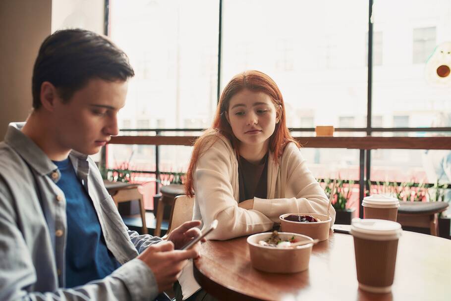 A woman with red hair sits at a table with food on it and looks at a man sitting next to her with a sad expression. The man looks down at a cell phone in his hand.