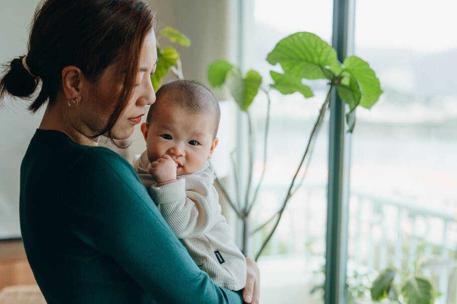 A mother in a green shirt holds her baby in her arms near a window as the baby looks at the camera.