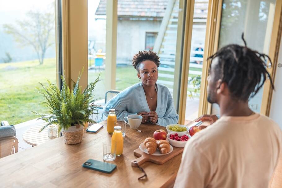 A male and female partner sit across from each other while eating breakfast at the kitchen table.