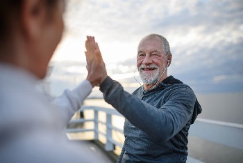 An elderly man in a blue long sleeve shirt and earphones is giving a high five and smiling to someone outside