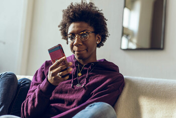 A young person with glasses and purple sweatshirt looks down at their phone with an unsure expression as they sit on the couch.