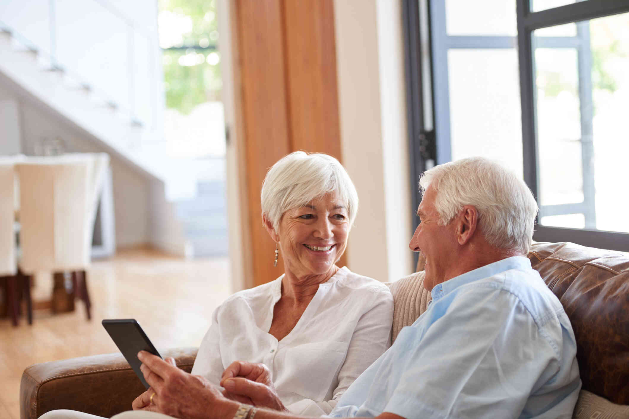 An elderly couple seated on a couch, sharing a warm moment as they look at each other, with the man holding a tablet.