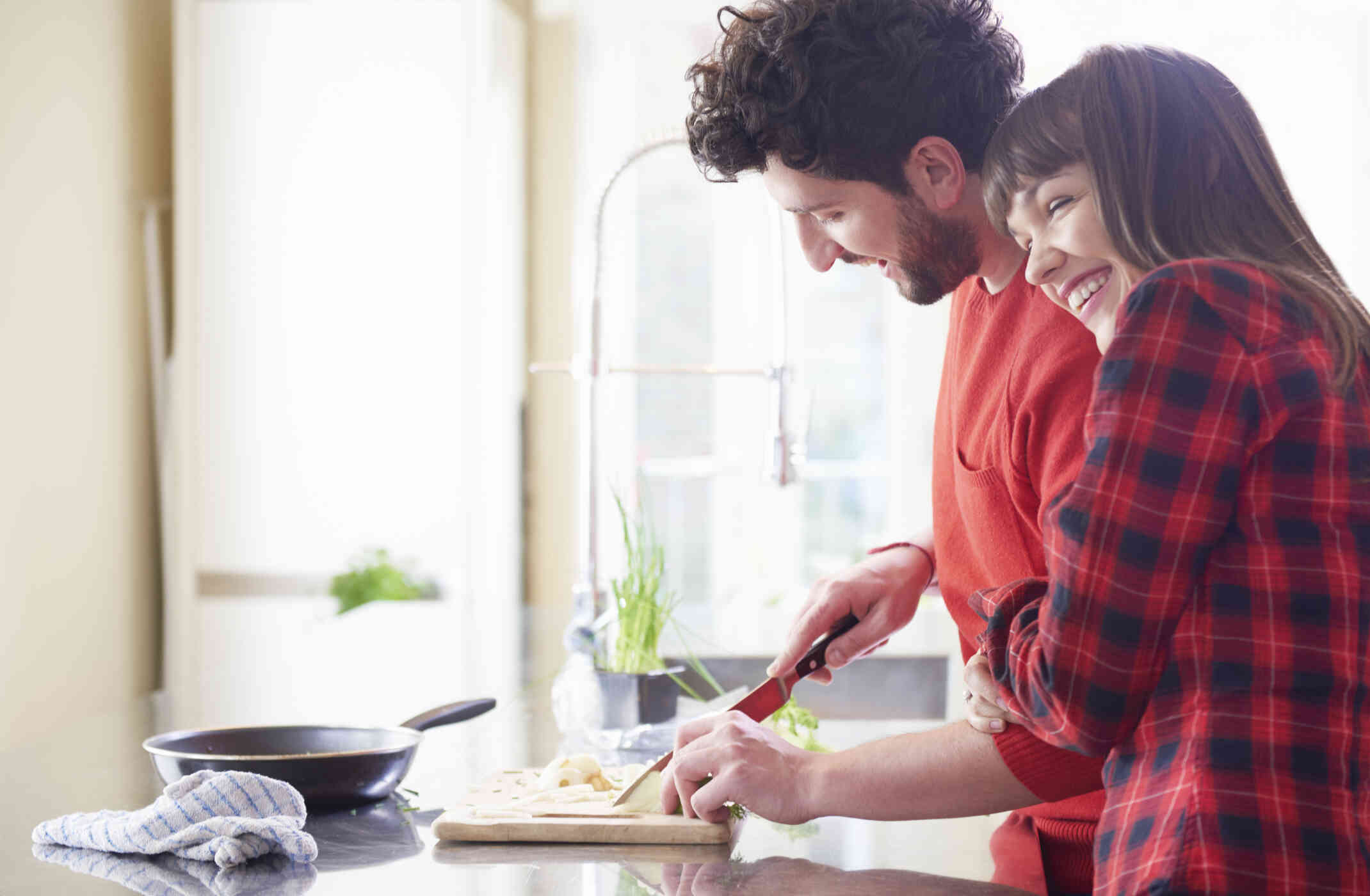 A man in a red shirt chops vegitables in the kitchen with a smile while his female partner gives him a hug.