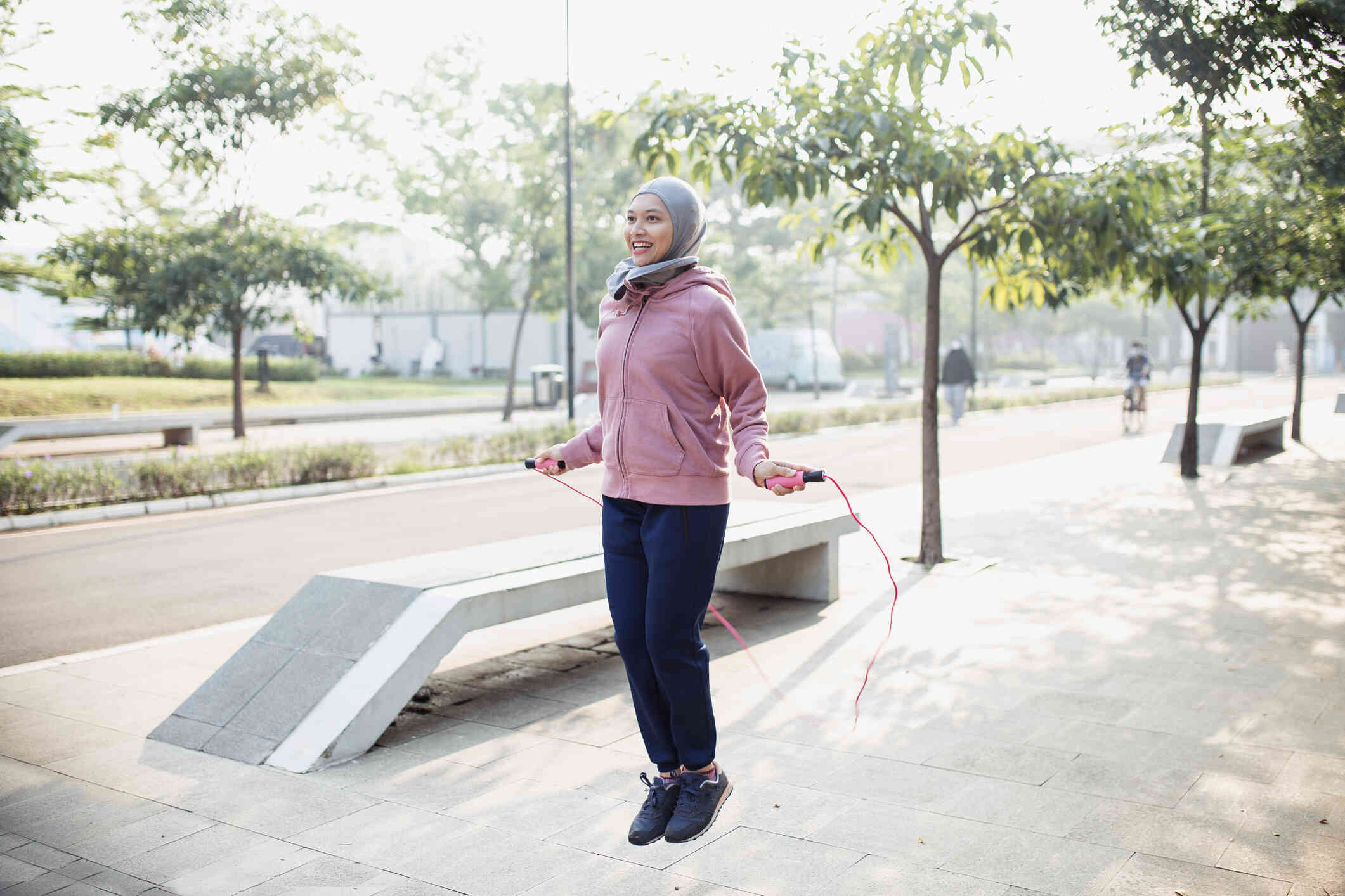 A woma in a hijab and pink sweater jumps rope while outsde on a sunny day.