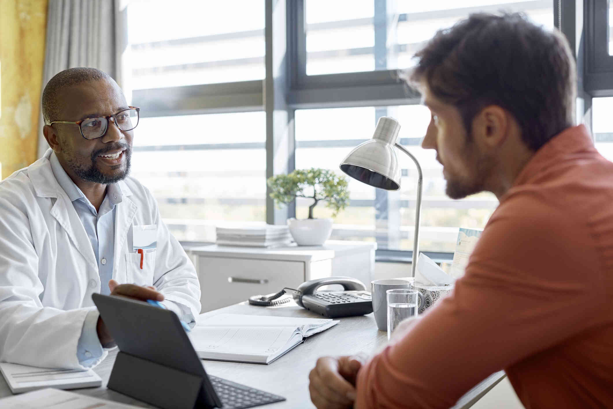 A man wearing a white coat and glasses sits behind a desk and speaks with another man who is sitting across from him in the office.