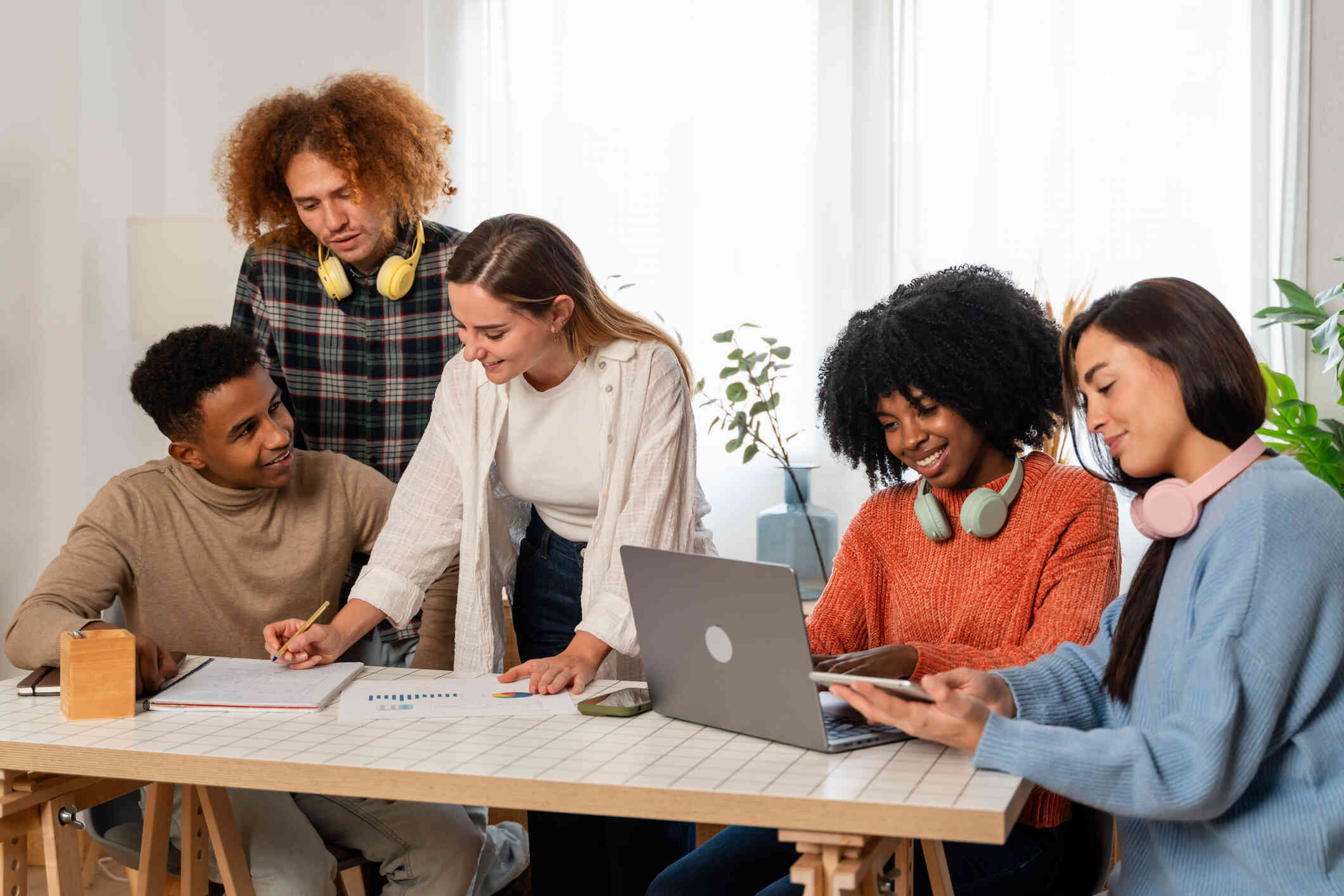 A group of teens sit around a desk while conversing and looking at a laptop with positive expressions