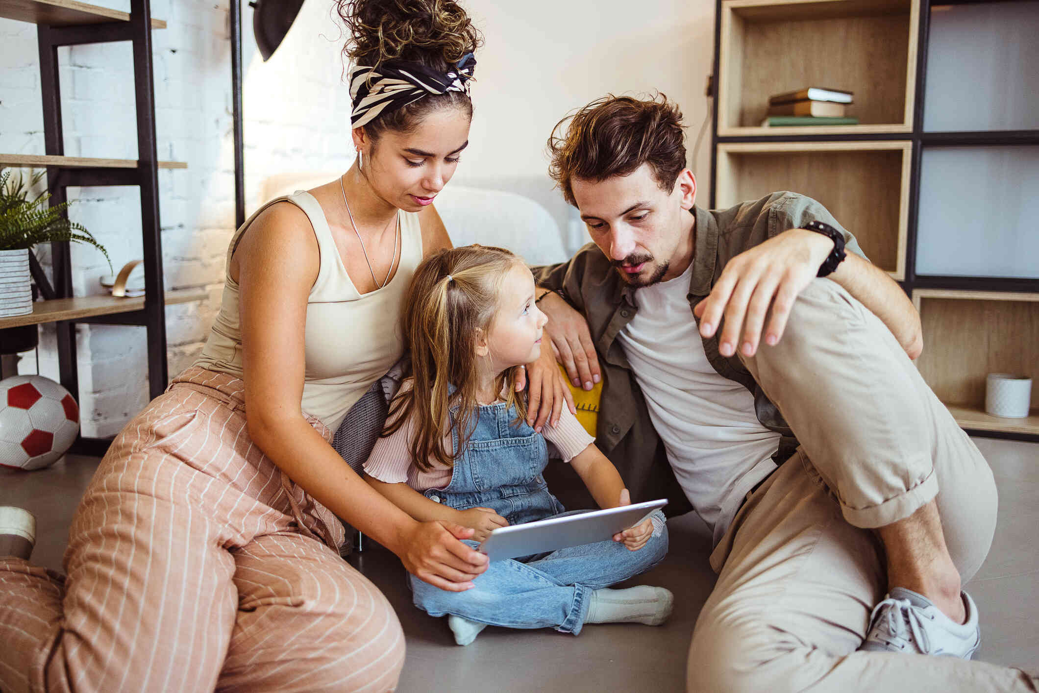 A family sits on the floor; the mother holds a tablet, the father speaks, and the child listens attentively.