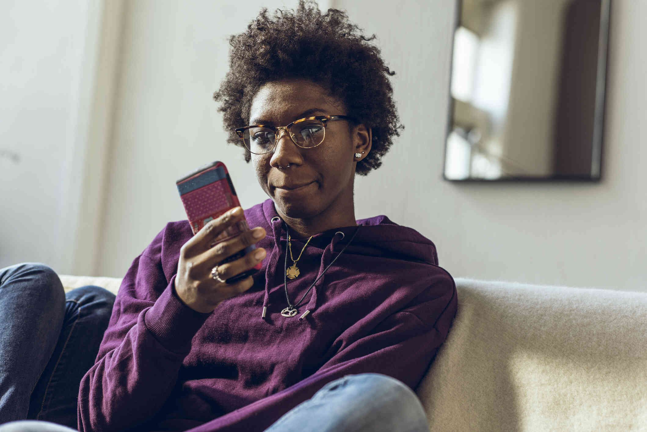 A woman in a purple hoodie sits on the couch and looks at the cellphone in her hand with  a curious expression.