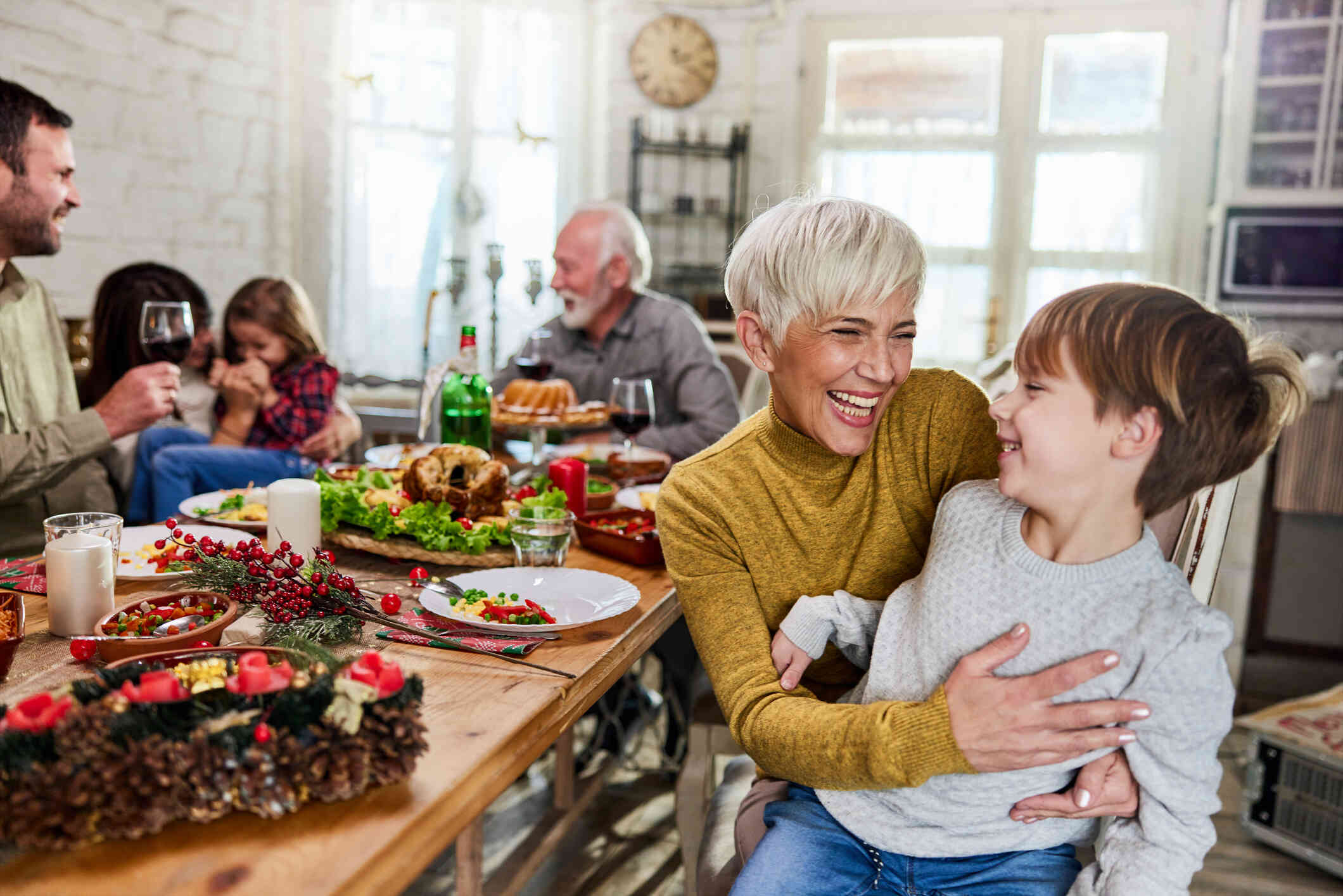 A woman with short, blonde hair smiles as she hugs a young boy. They are sitting at a table with other people and the table is filled with food.