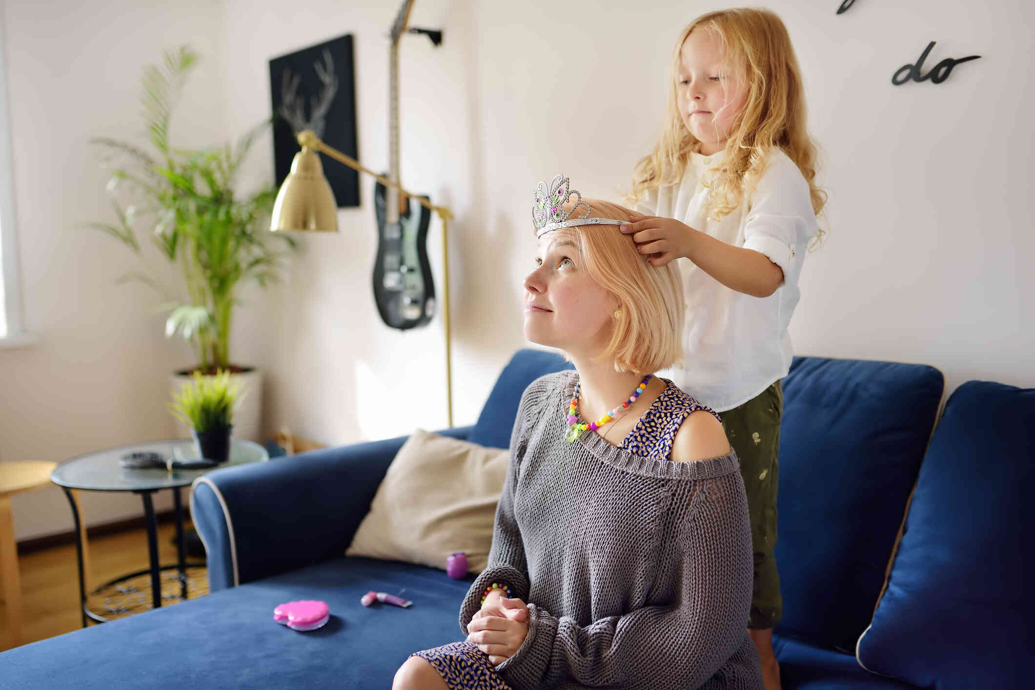 A mother sits on the couch as her daughter stends behind her and places a plastic tiara on her mothers head.