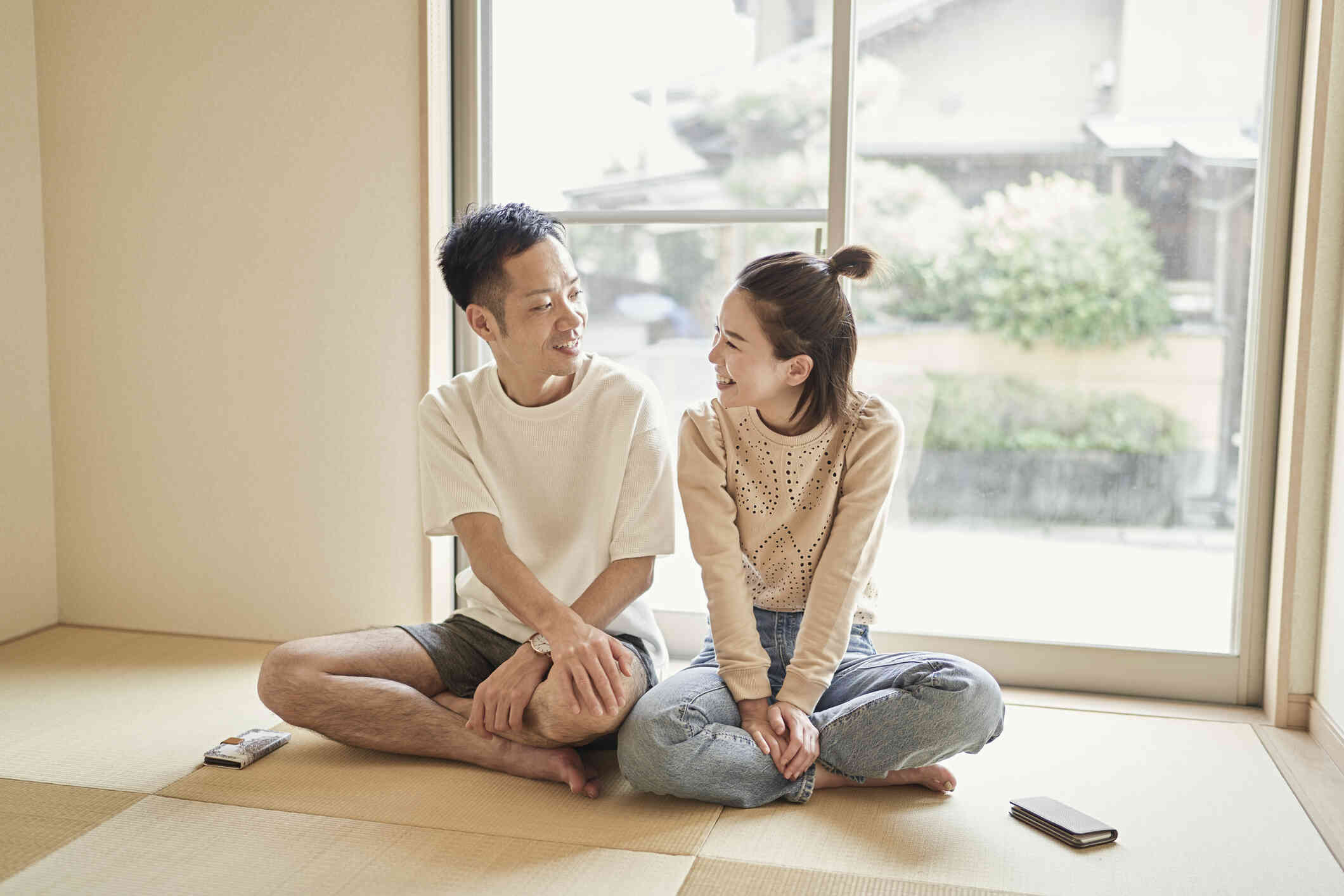 A male and female couple sit side by side cross legged on the florr while smiling and talking.