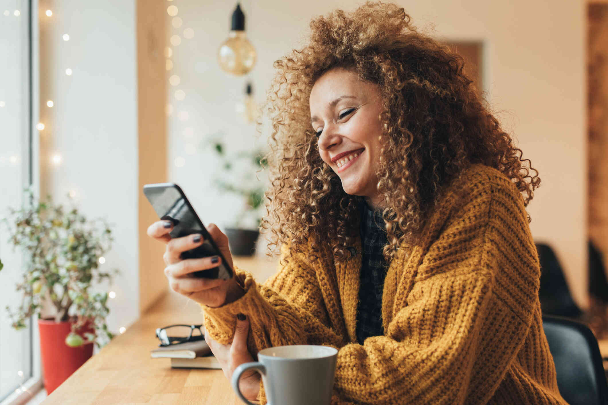 A woman in a yellow sweater sits at a coucnter with her coffee cup and smiles brightly while looking at the cellphone in her hand.