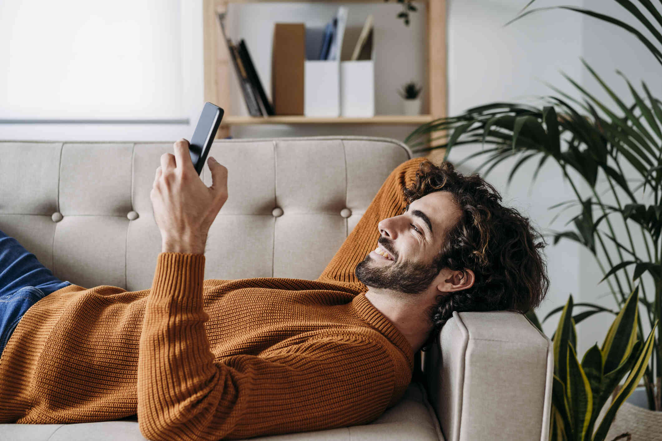 A man in an orange sweater lays on his back on the couch and smiles while looking at the phone in his hand.