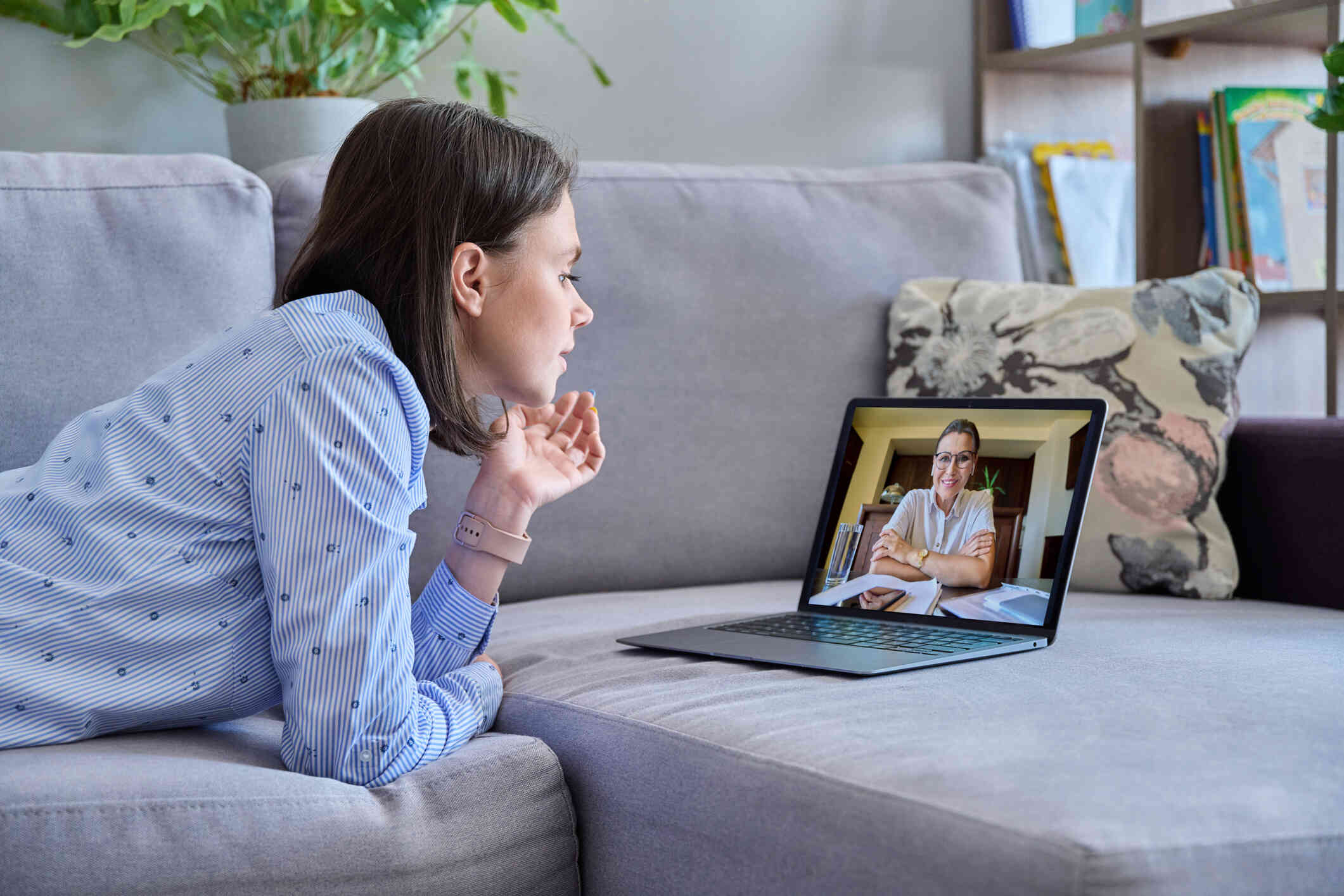 A young woman in a blue shirt lays on couch while talking with an online therapist on the computer