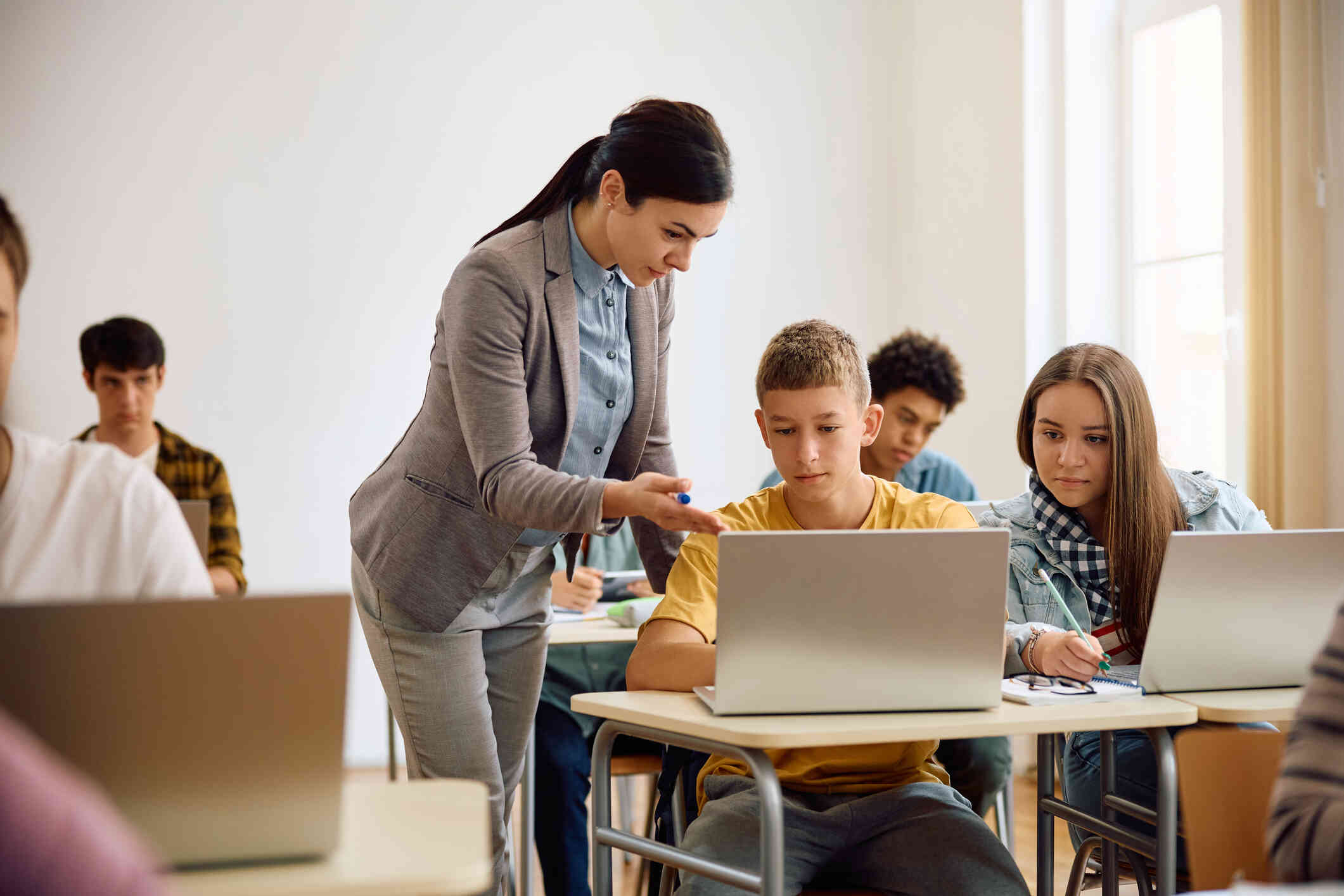 A teacher helps a young boy with his laptop while a classmate watches attentively.