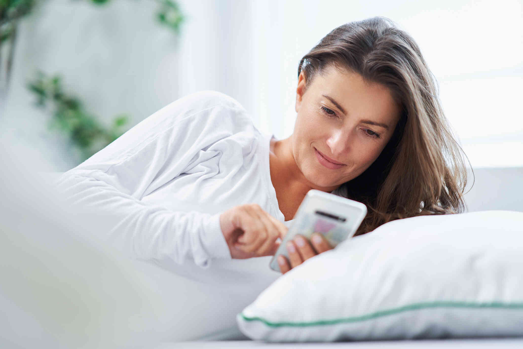 A woman in a white shirt reclines in bed and loks at the cellphone in her hand.