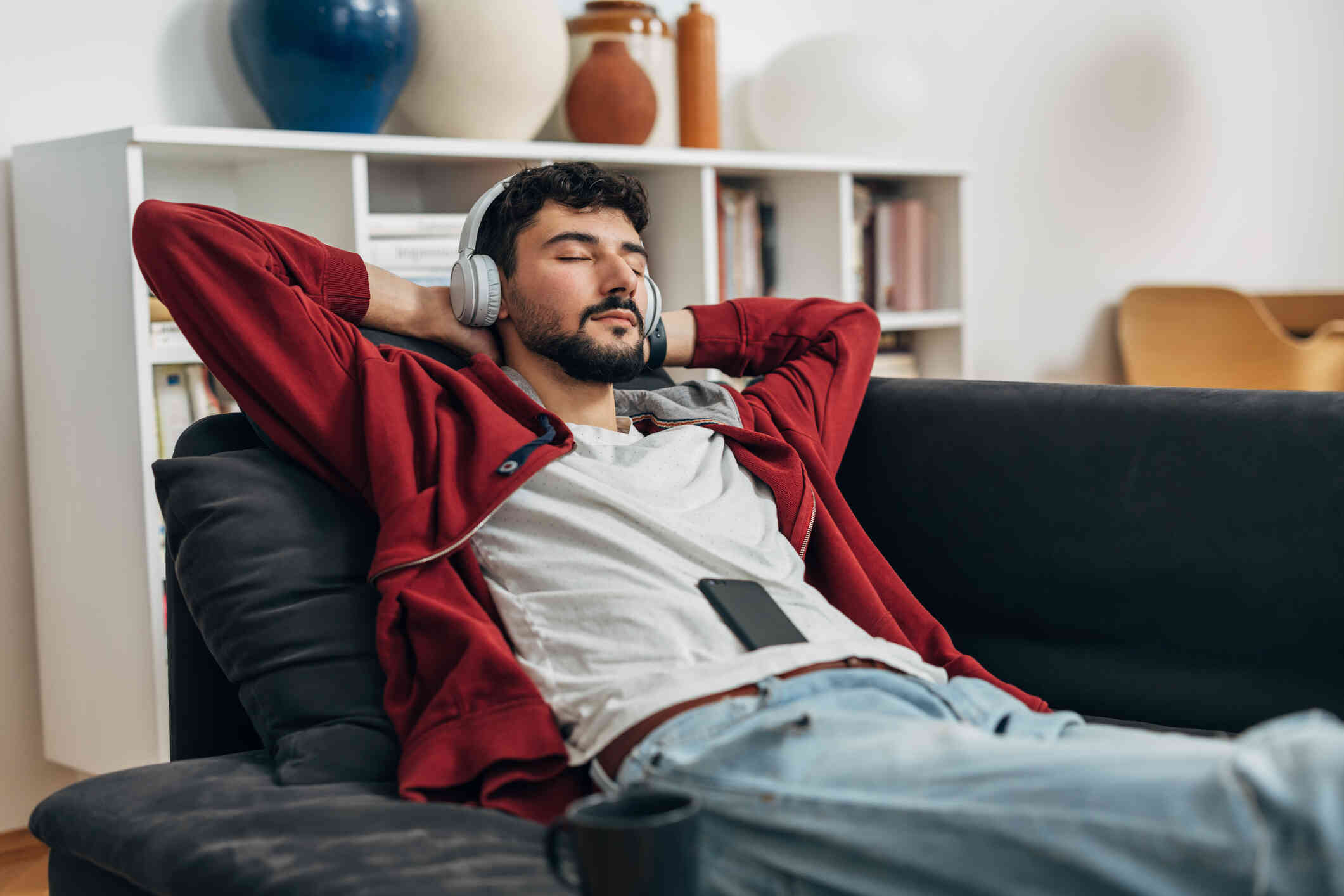 A man reclines on the couch with his eyes closed and  headphones on as his cellphone rests on his stomach.