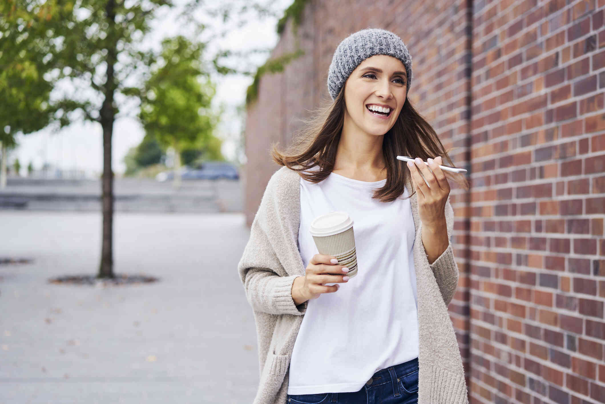 A woman in a beaning holds a cup of to go coffee in one hand and her phone in the other while talking on the phone and smiling as she walks down the sidewalk.