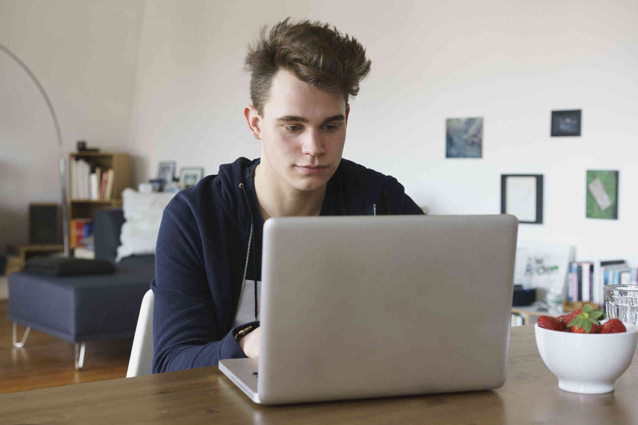 A teen boy sits in a chair ant a table behind a laptop and looks at the screen with a serious expression.
