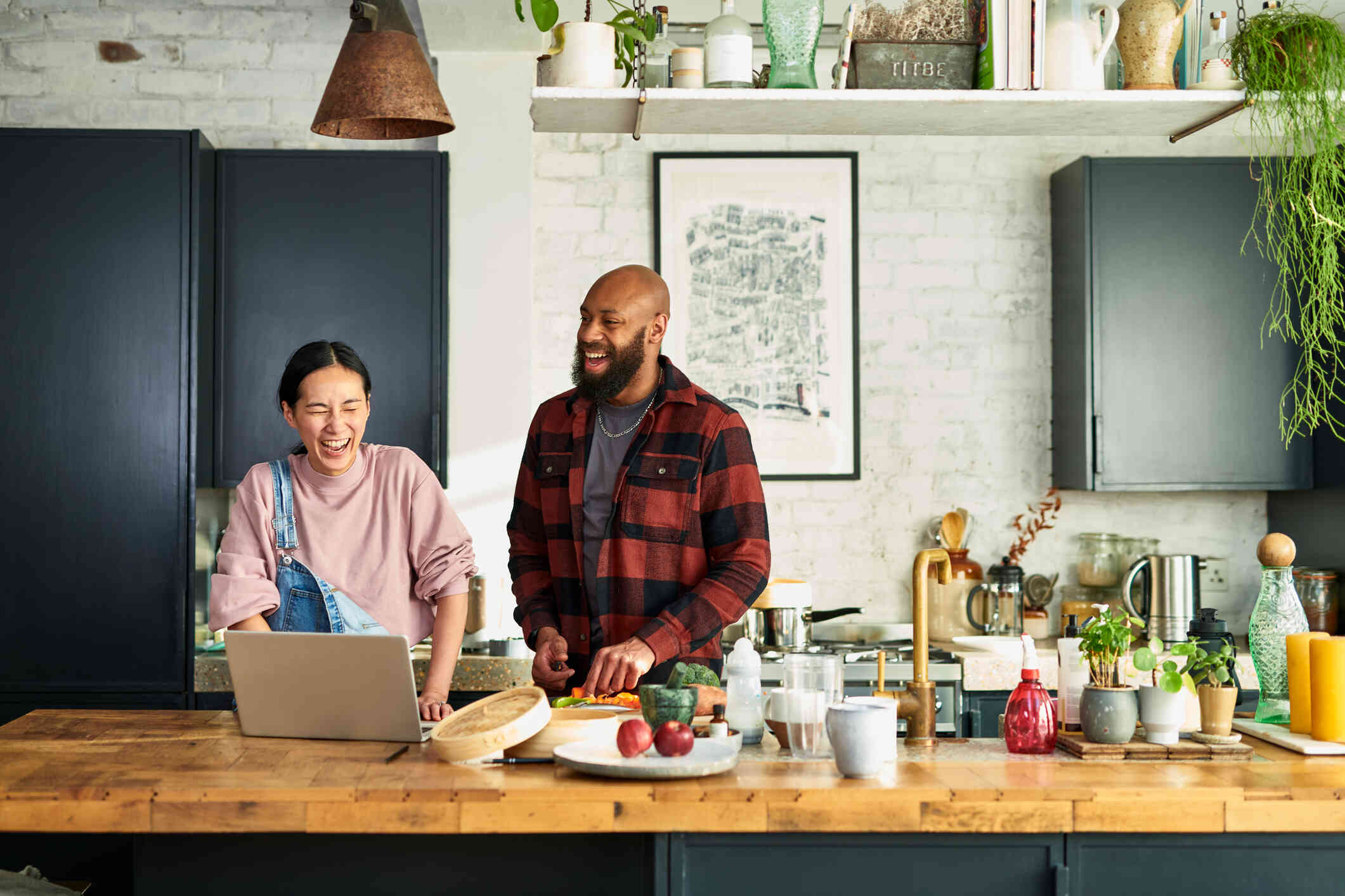 A woman in overalls laughs as she stands next to a man in a red plaid shirt who is chopping vegetables in the kitchen.