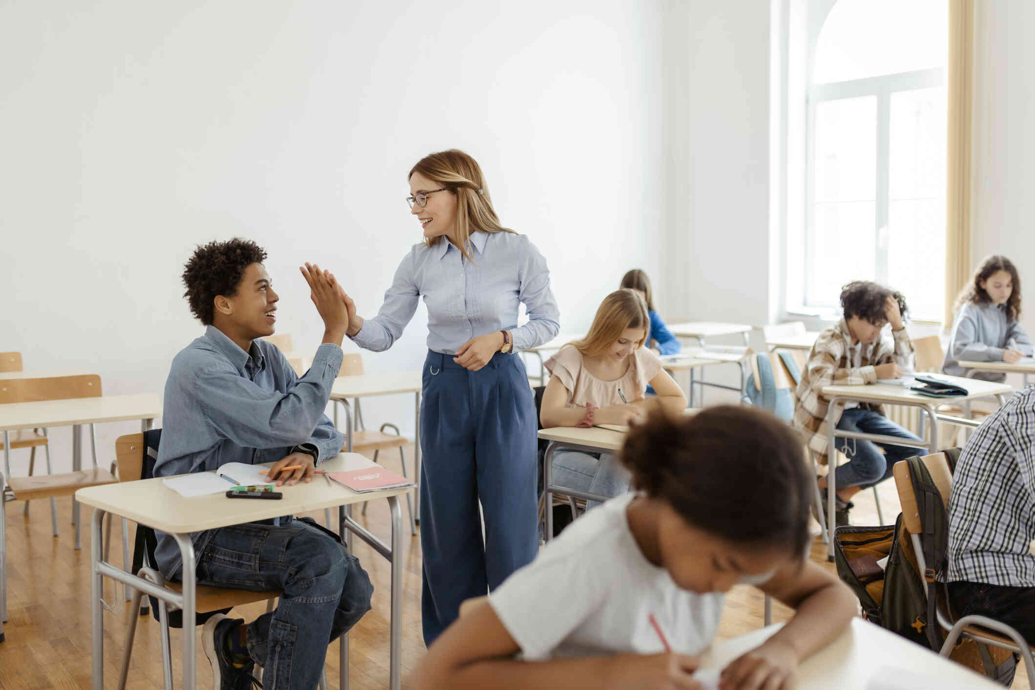 A teenage boy in a blue shirt sits in a desk in a classroom while high-fiving a teacher standing next to him as they both smile