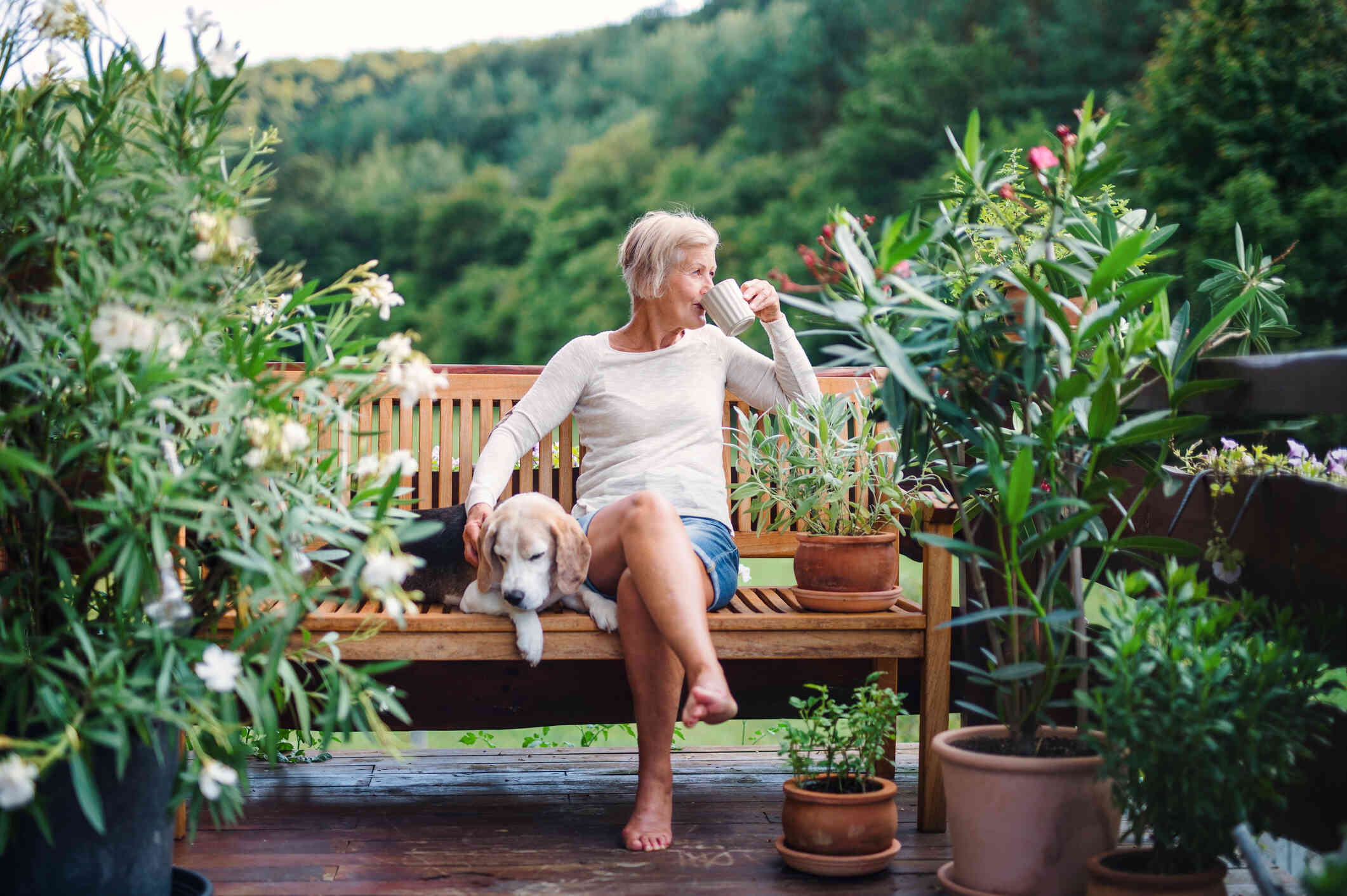 A mature woman sits on a bech outside with her dog as she sits from a cup of coffee.