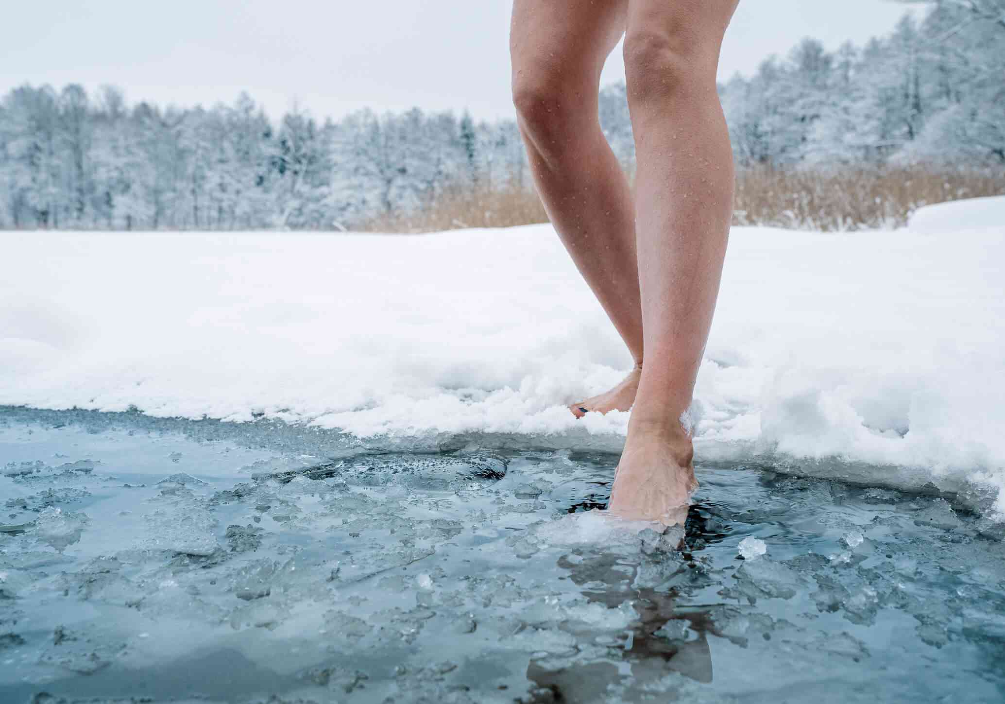 A close up of a woman dipping her bare feet into the cold water of a pond while surrounded by snow.