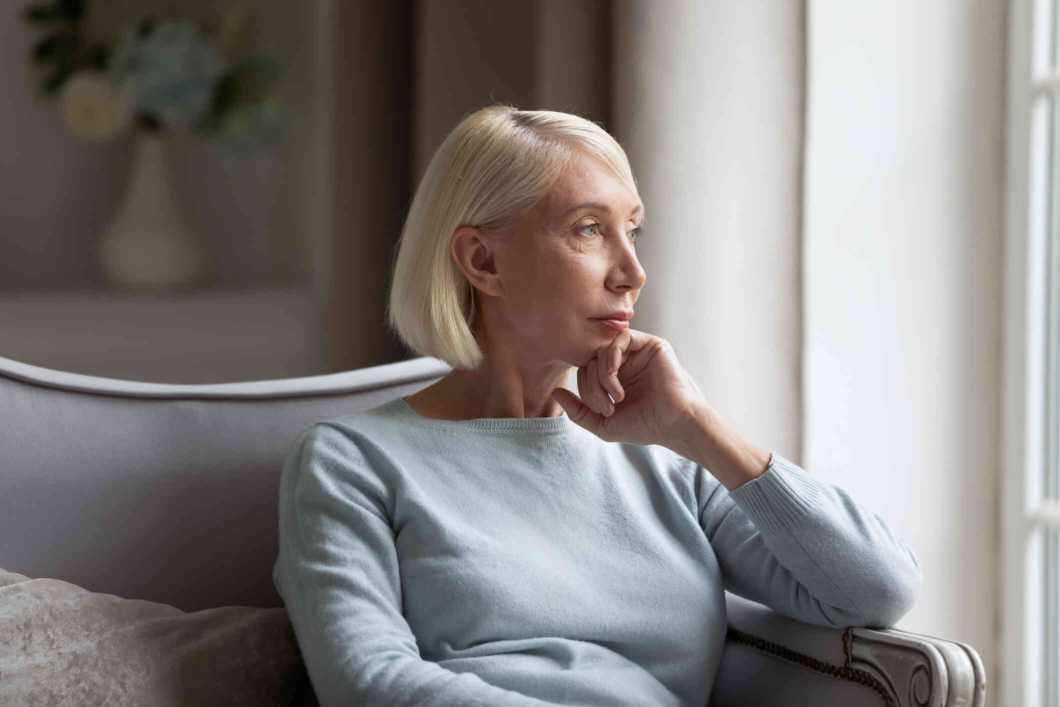 A middle aged woman sits in an armchair and rests her chin against her hand while gazinf off deep in thought.