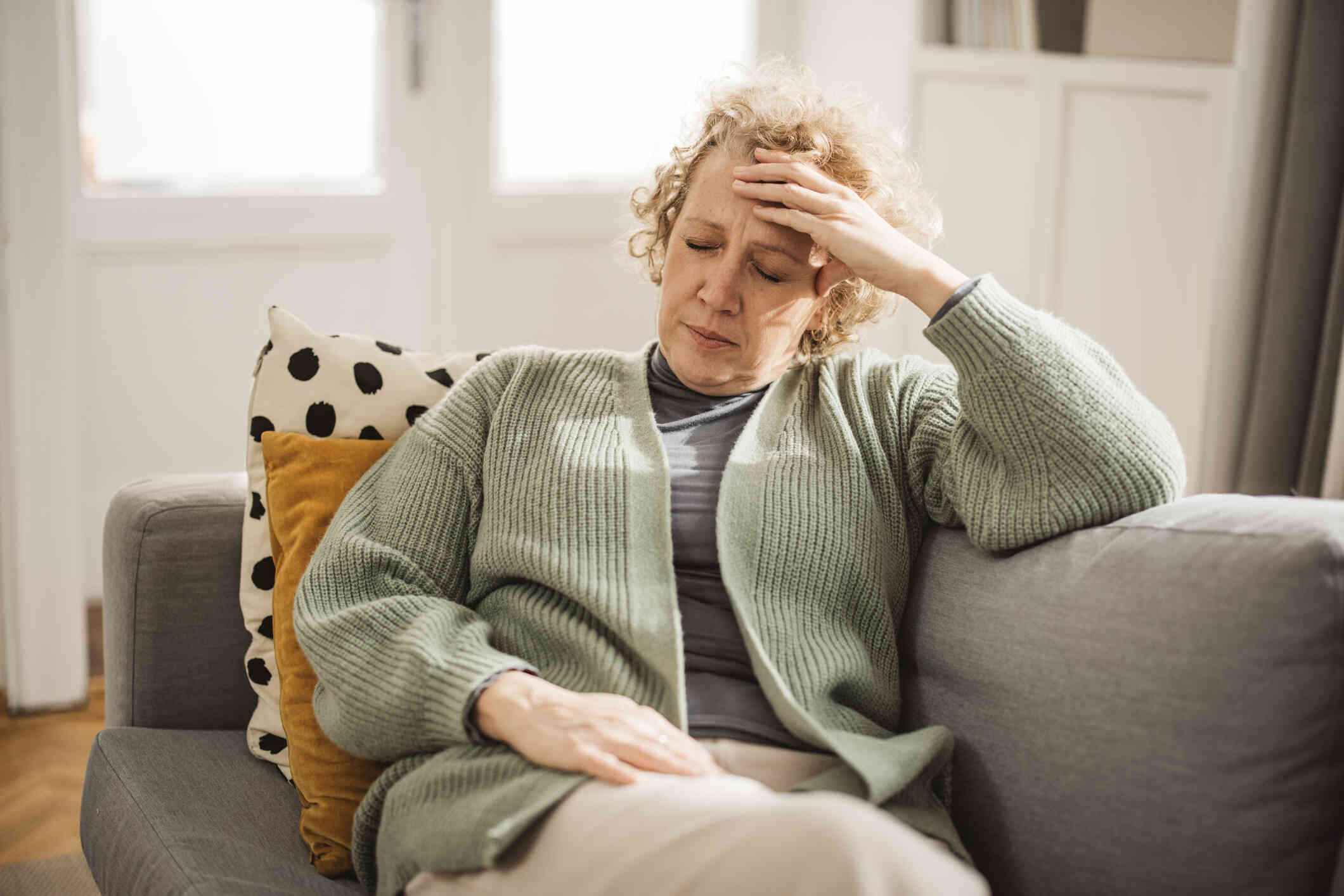 A mature woman in a green sweater sits on her couch with her head resting sadly in her hand and her eyes closed.