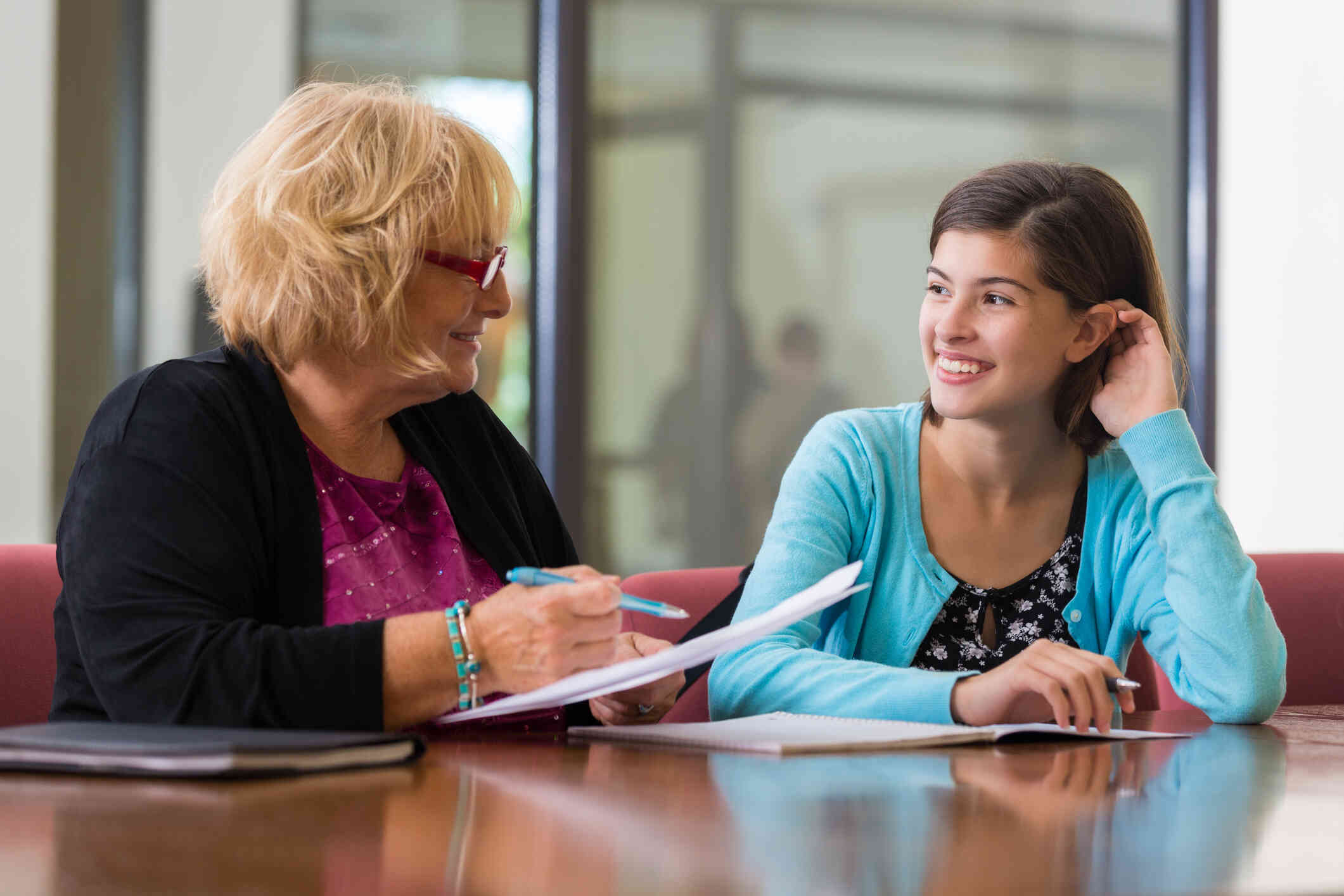 A mature woman with short hair and glasses holds a pen and paper as she sits at a table and speaks to a young woman who is sitting next to her smiling.