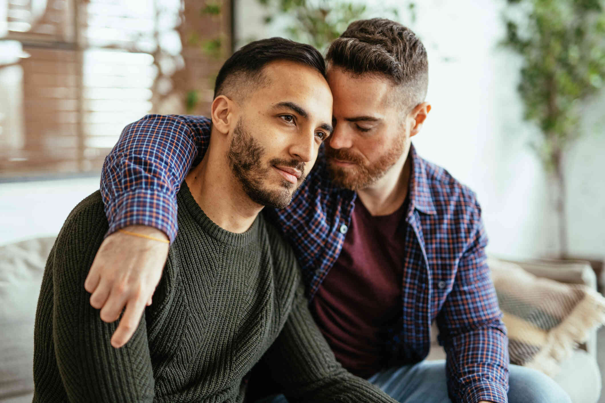 A man wraps his arm around his male partner as they sit next to each other with worried expression.