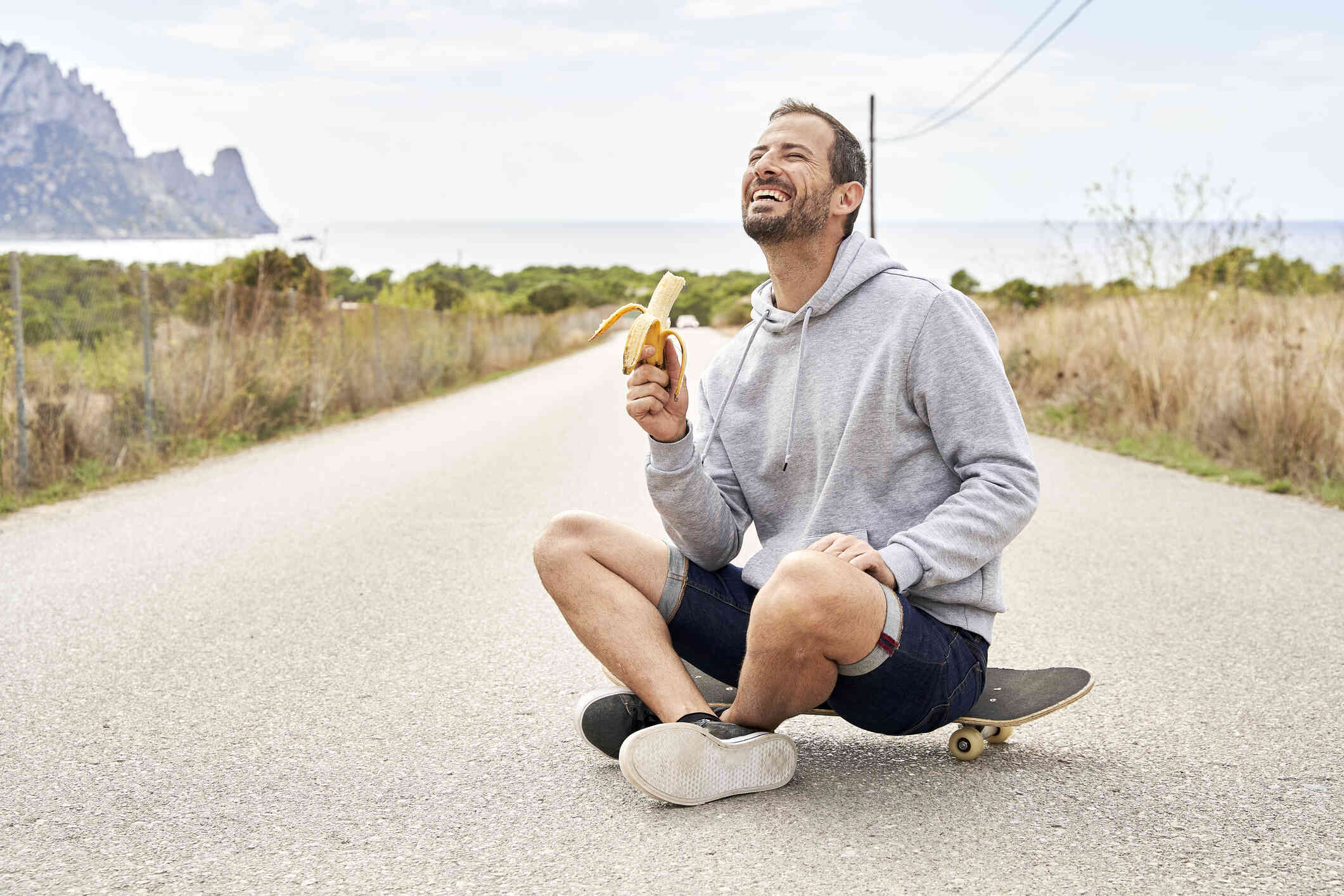 A middle aged man in a grey sweater sits on a skaptboard while holding a half eaten banana and smiling while looking off.