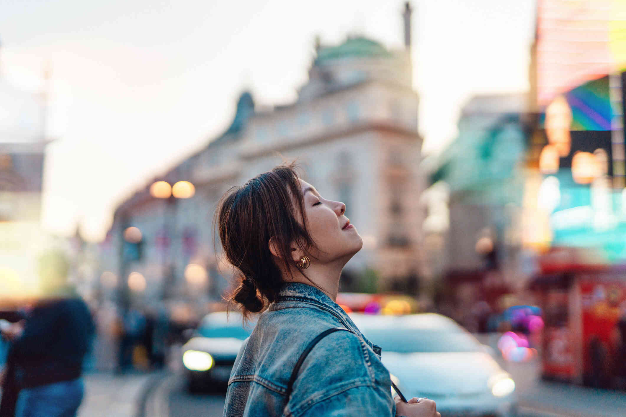 A woman stands outside near traffic in a busy city and closes her eyes while lifting her head up.