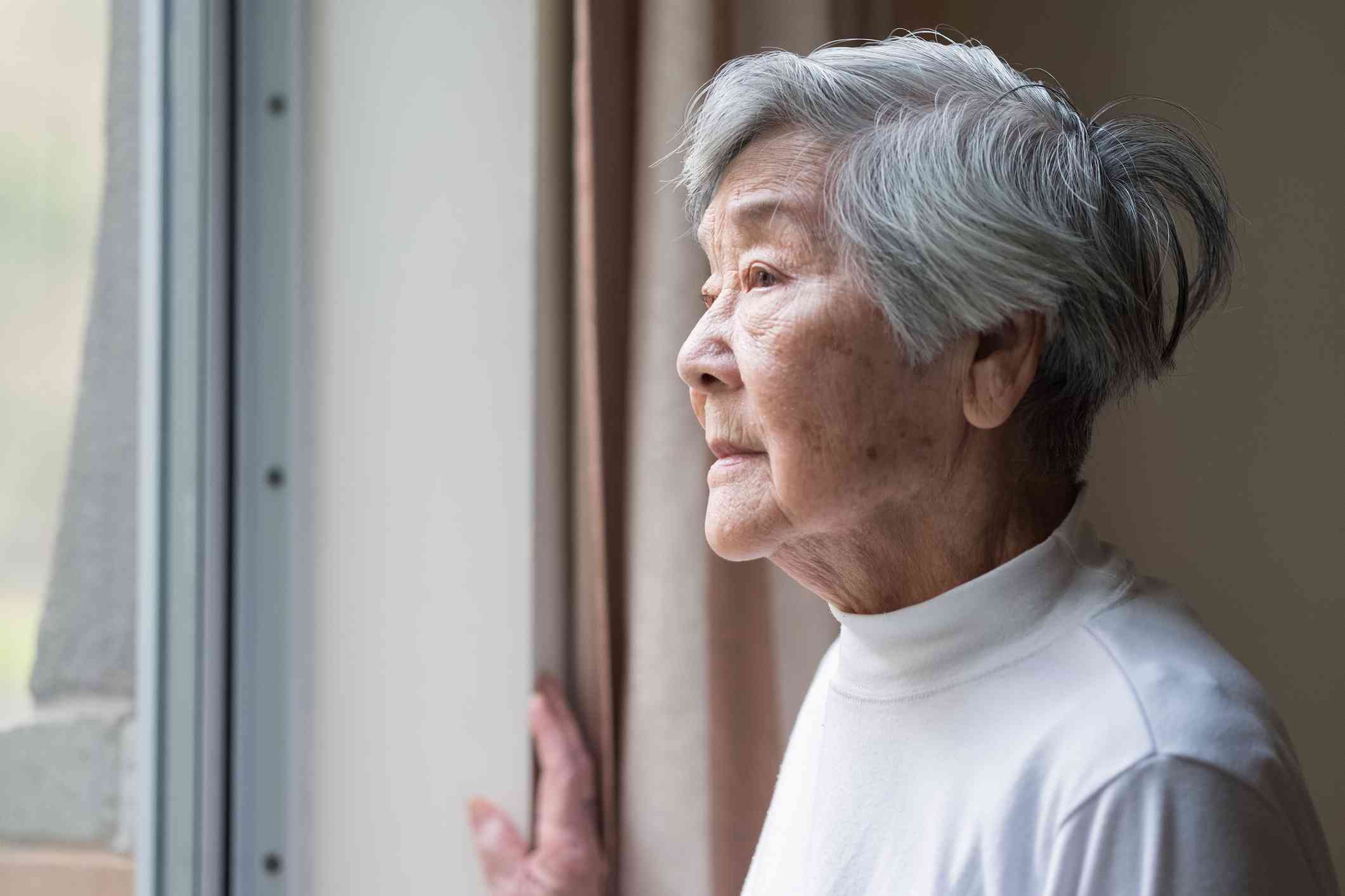 An elderly woman in a white shirt stands near a window in her home and gazes out sadly.
